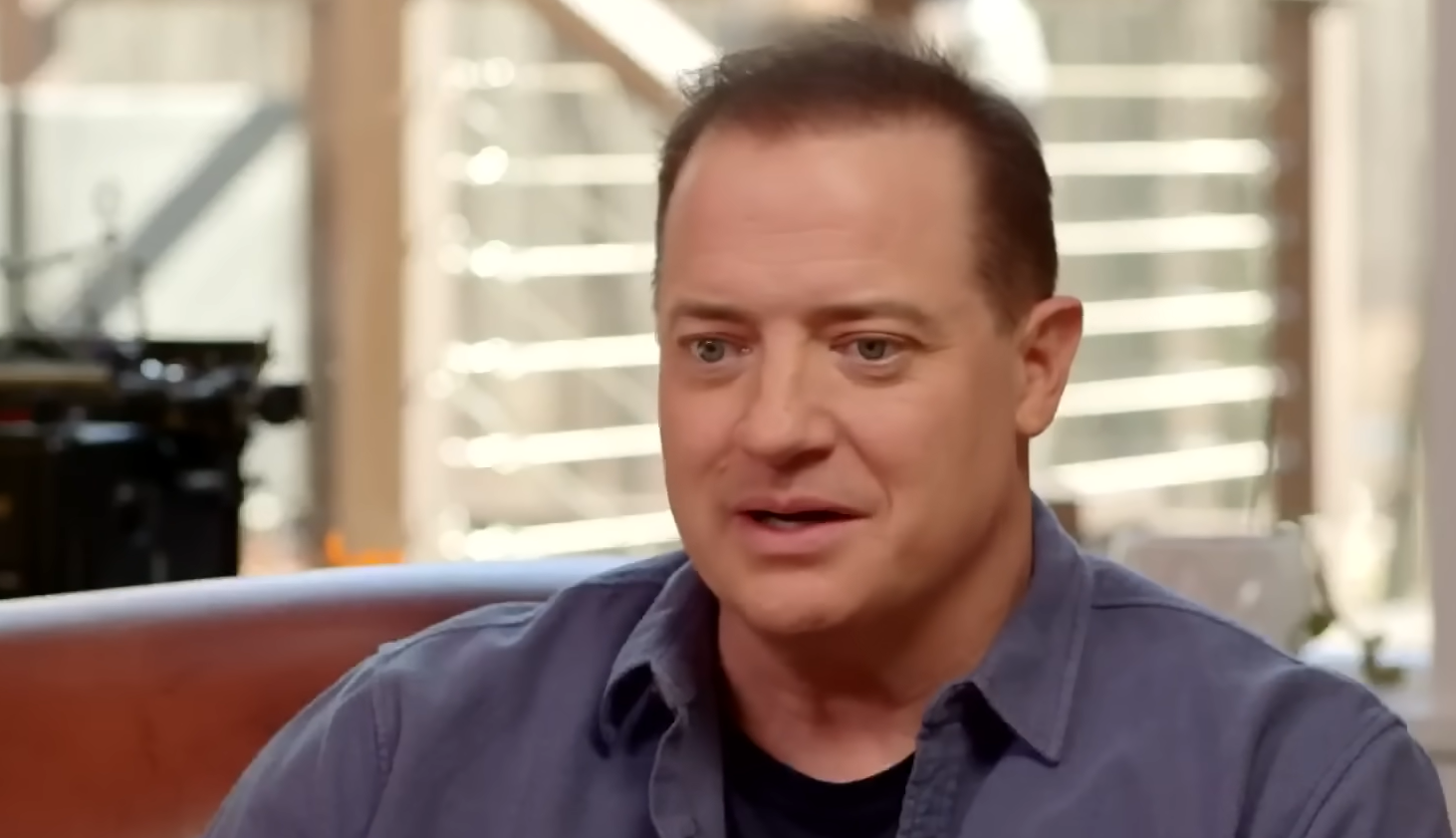 Brendan Fraser in a blue shirt, sitting on sofa in front of the camera in the room