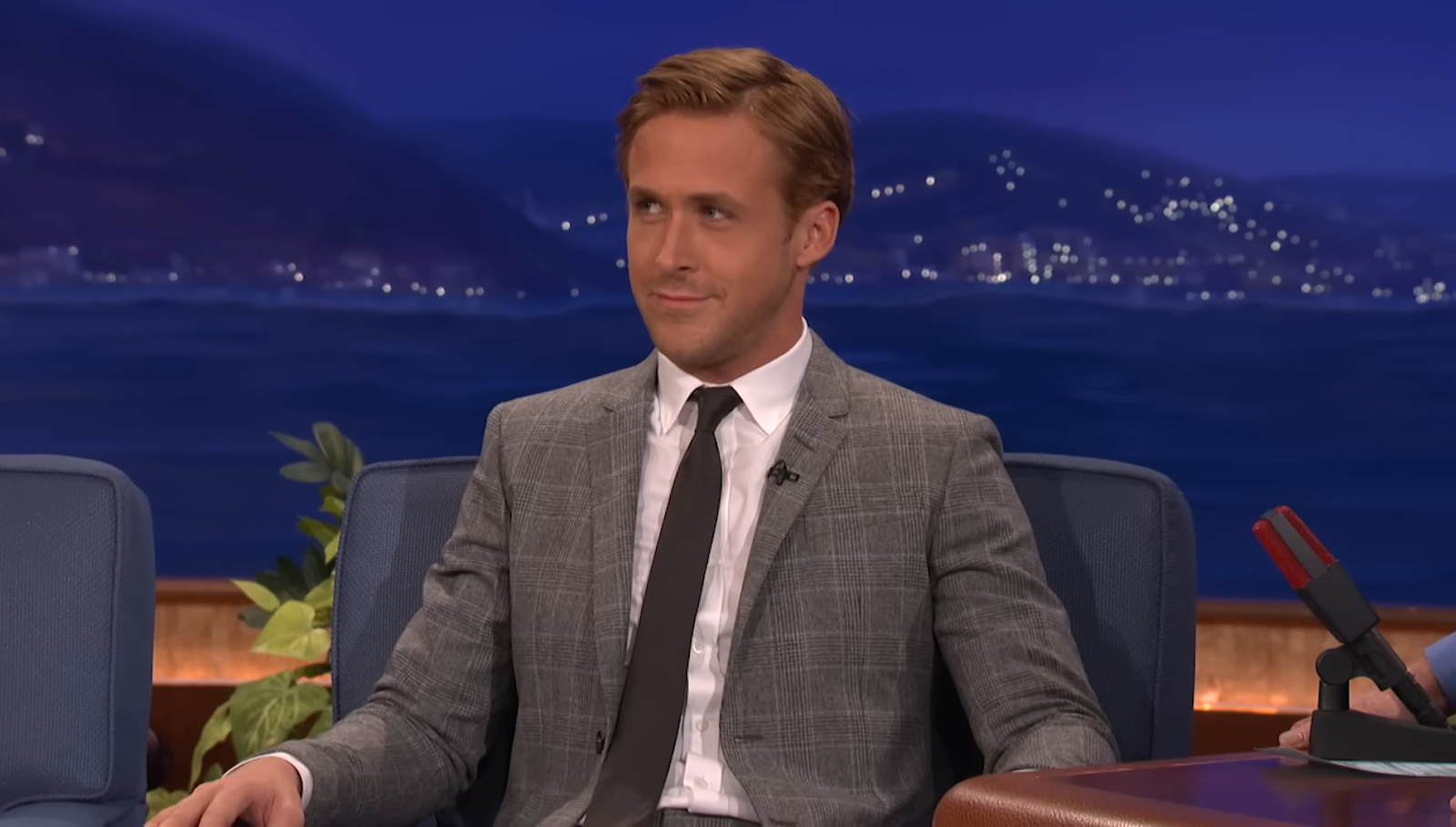 Ryan Gosling in a gray classic suit and tie sitting in a chair in front of the city view behind him