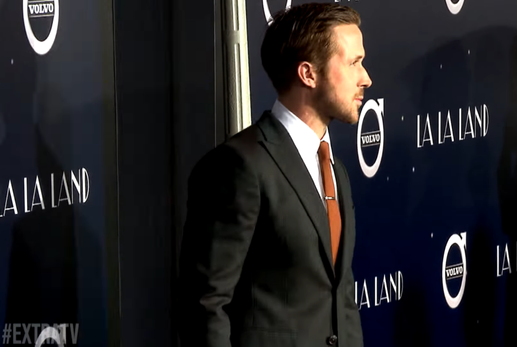 a side view of Ryan Gosling in a black classic suit and brown tie standing near the wall with Lalaland written on it