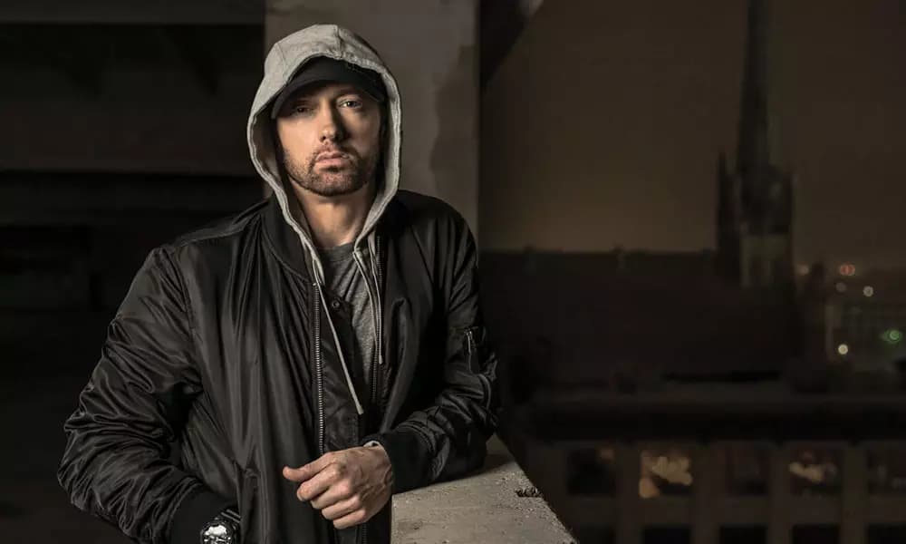 Eminem stands on a balcony wearing a hoodie and looking at the camera