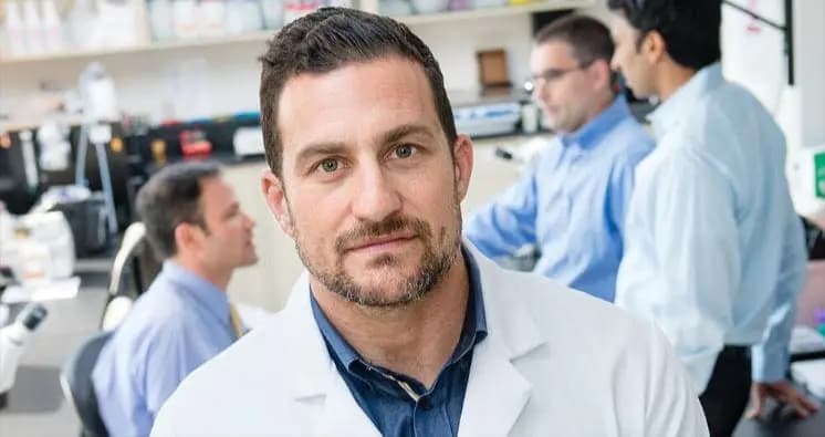 A man with a beard stands in a white coat